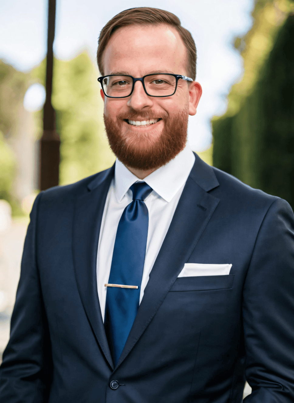Man with glasses and a beard wearing a suit and blue tie, smiling outdoors.