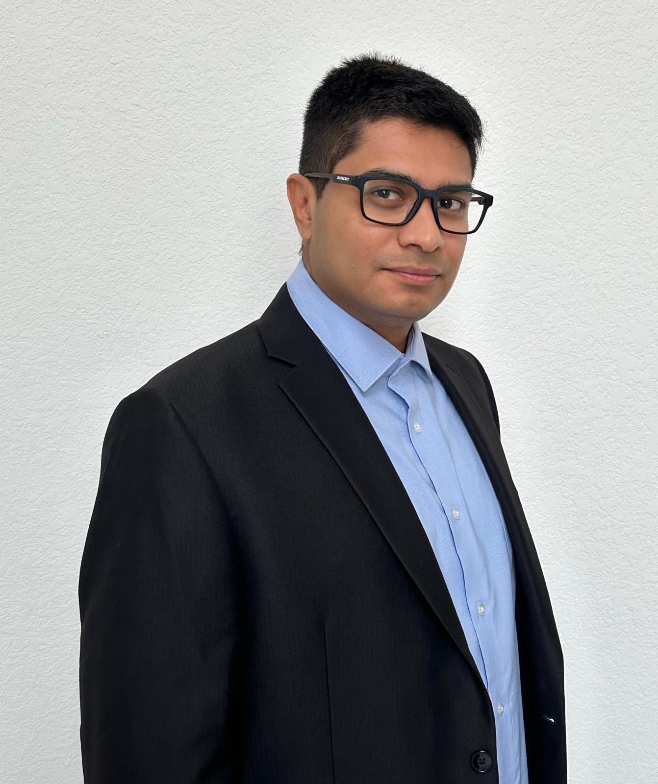 Man wearing a blue collared shirt and black blazer, standing against a white textured wall.